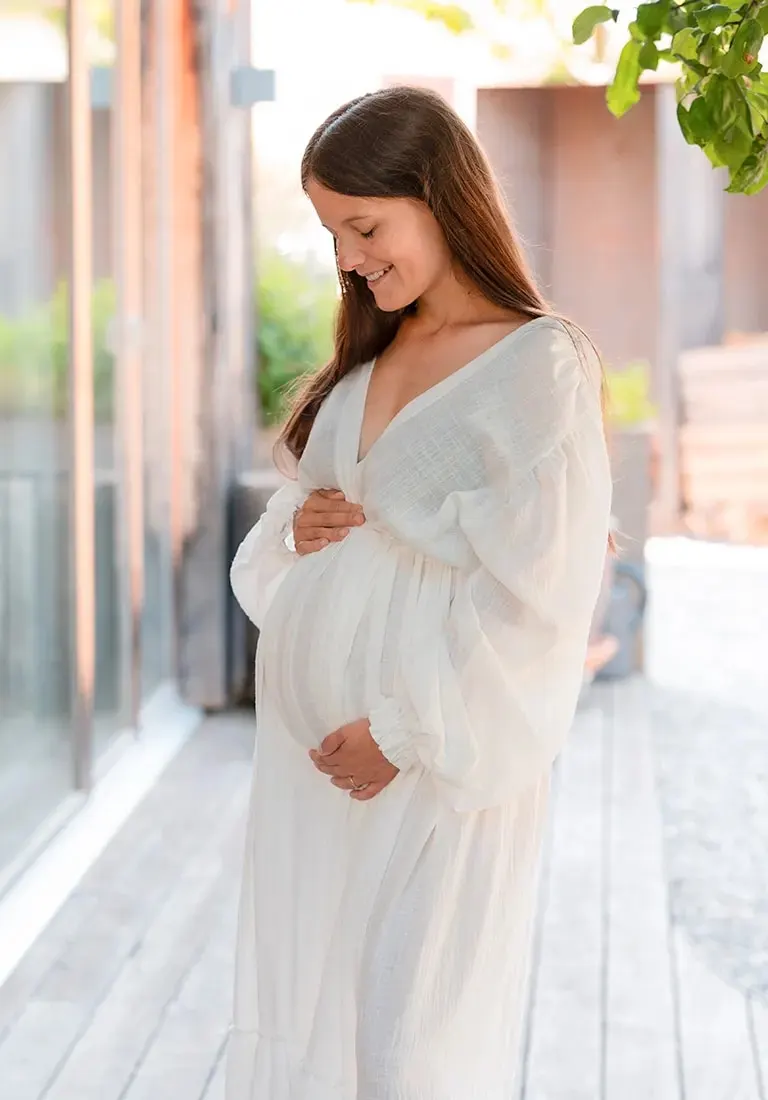 Schwangere Frau in einem weissen Kleid, die lächelnd ihren Bauch hält