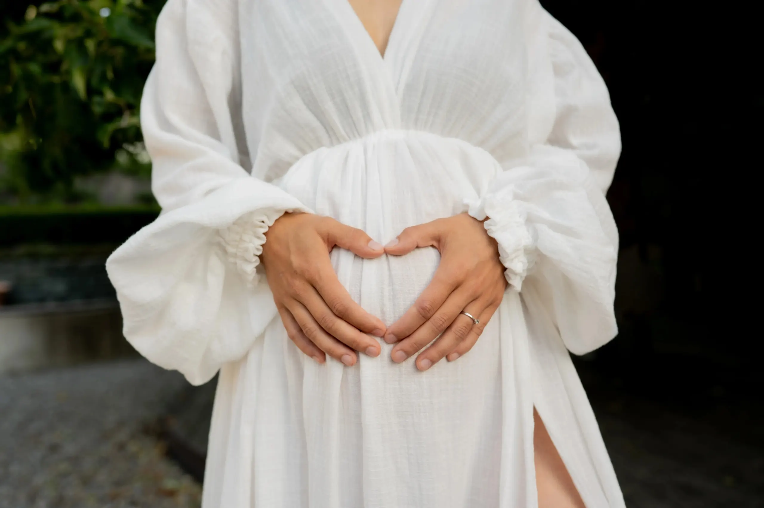 Schwangere Frau in einem weissen Kleid formt mit ihren Händen ein Herz auf dem Bauch