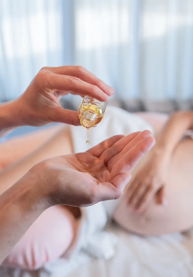 Massageöl wird in eine Handfläche gegossen, dahinter liegt eine schwangere Frau auf der Massageliege